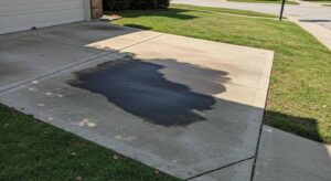 A suburban concrete driveway with a large dark oil stain, bordered by a patch of grass and part of a house in the background.